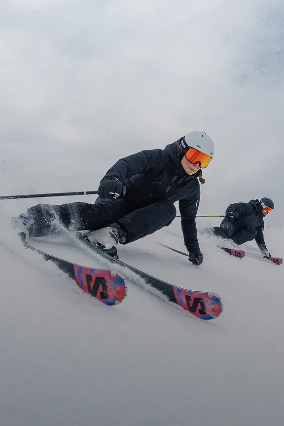 Zwei Skifahrer in dunklen Anzügen carven dynamisch einen verschneiten Hang hinab; der vordere ist im Fokus.