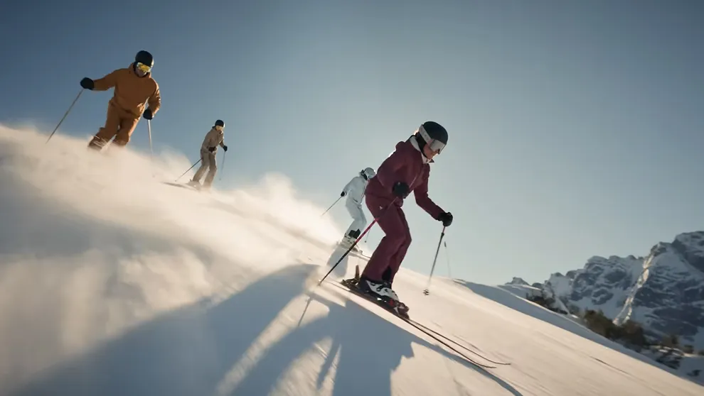 Vier Skifahrer auf sonniger Piste vor blauem Himmel. Im Fokus carvt eine Person in weinrotem Anzug.