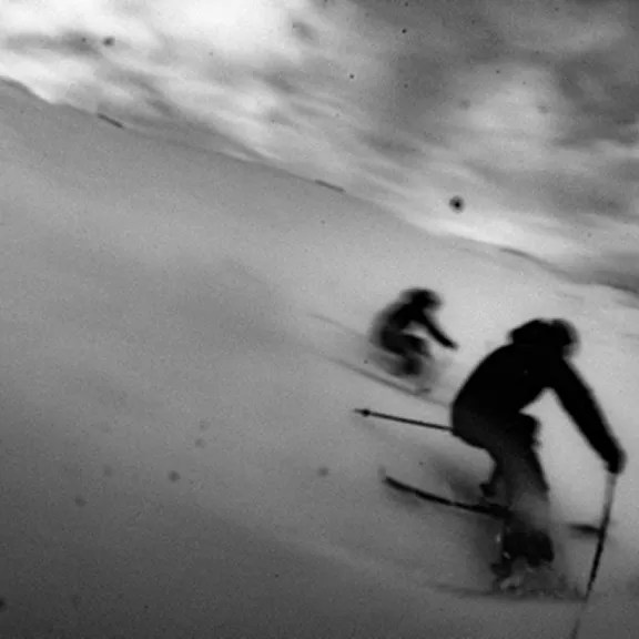 Körniges Schwarz-Weiß-Foto: Zwei Skifahrer rasen dynamisch den Hang hinab. Bewegungsunschärfe vermittelt hohes Tempo.